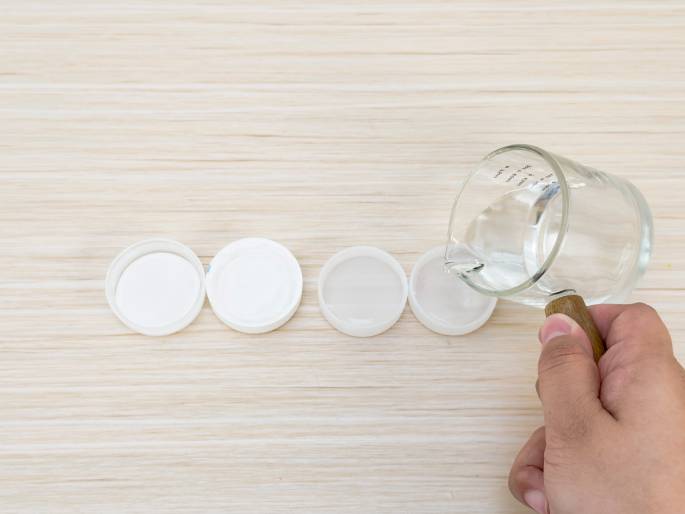 A hand pours clear liquid from a small glass measuring cup with a wooden handle onto a row of four white plastic bottle caps arranged on a light woodgrain table. The image suggests an experiment or activity involving precise measurement of liquids.