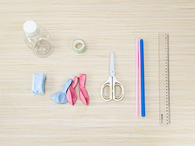 An overhead flatlay on a light woodgrain surface displays various items, likely for a science experiment or craft project. These include an empty clear plastic bottle lying on its side, a roll of clear tape, a small blue package (possibly containing powder or a small object), two deflated balloons (one light pink and one light blue), a pair of white-handled scissors, two straws (one pink and one blue), and a clear plastic ruler. The arrangement suggests preparation for an activity involving these materials.