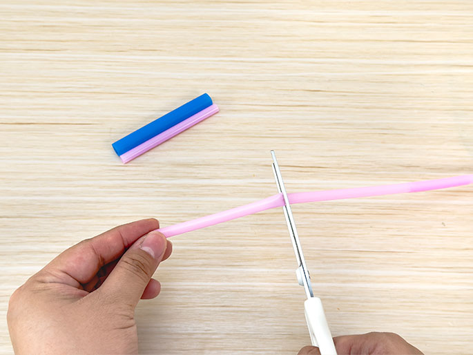 A pair of hands uses scissors to cut a pink flexible straw against a light woodgrain background. A shorter, already-cut piece of pink and blue straw lies near the top of the frame. This image likely demonstrates a step in a DIY project or science experiment involving the straws.