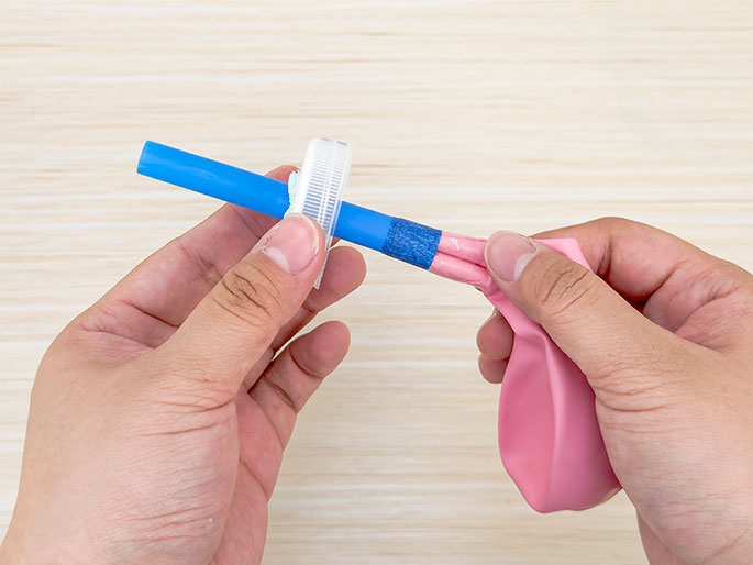 Two hands are shown against a light woodgrain background. One hand holds a blue straw which has a white plastic bottle cap attached to one end. The other hand holds a deflated pink balloon, the neck of which is stretched and taped around the other end of the blue straw. This image likely depicts the construction of a simple balloon-powered projectile launcher or a science experiment demonstrating air pressure.
