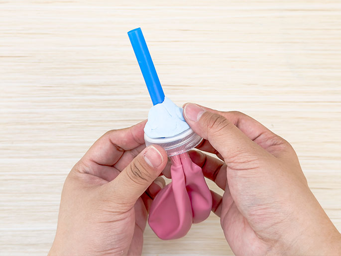 Two hands hold a small device constructed from a plastic bottle cap, a blue straw, and a pink balloon. Light blue putty or clay seals the area where the straw enters the bottle cap. The neck of the deflated pink balloon is attached to the bottom of the bottle cap. The background is a light woodgrain surface. This image likely depicts a homemade toy or a science experiment, possibly a balloon-powered pump or projectile launcher.