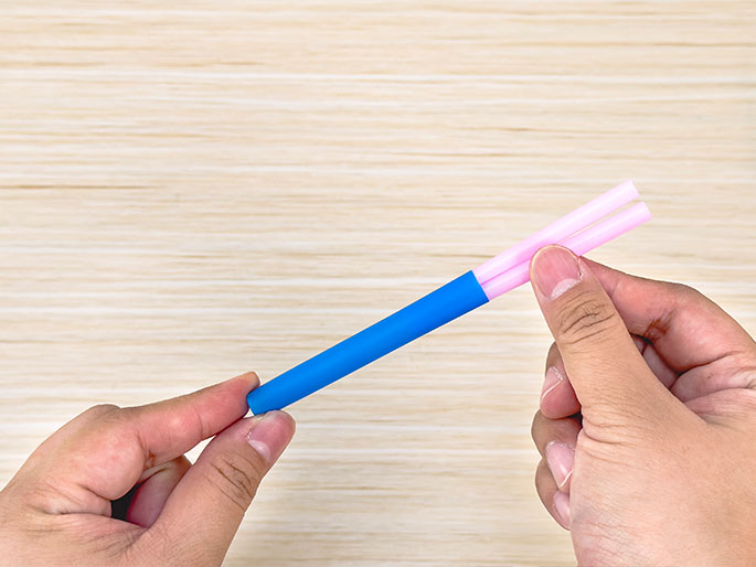 Two hands hold a blue and pink straw against a light woodgrain background. A shorter section of a pink straw is inserted into the end of the blue straw. This close-up image likely illustrates a step in constructing something using straws, possibly for a craft or science experiment.