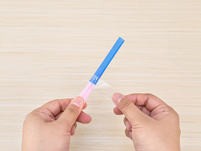 Two hands hold a blue and pink straw with a piece of clear tape being wrapped around the joint where the two straws meet, against a light woodgrain background. This close-up image likely illustrates a step in a DIY or science project where the straws are being connected securely.