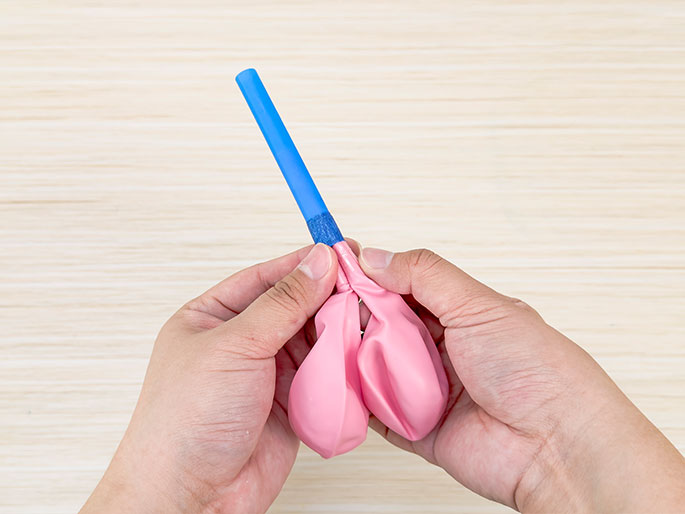 Against a light woodgrain background, two hands are shown attaching a deflated pink balloon to a blue straw. The straw, secured with tape at the connection point, is inserted into the neck of the balloon, which is being held and pinched to create a seal. This image likely demonstrates a step in a science experiment or a DIY project involving balloons and straws.
