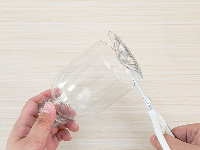Against a light woodgrain background, two hands are shown cutting a clear plastic bottle with a pair of scissors. The bottle has already been partially cut, and the top portion, which includes the bottle's neck and cap, is being trimmed further. This image likely depicts a step in a DIY project or craft activity involving repurposing a plastic bottle.