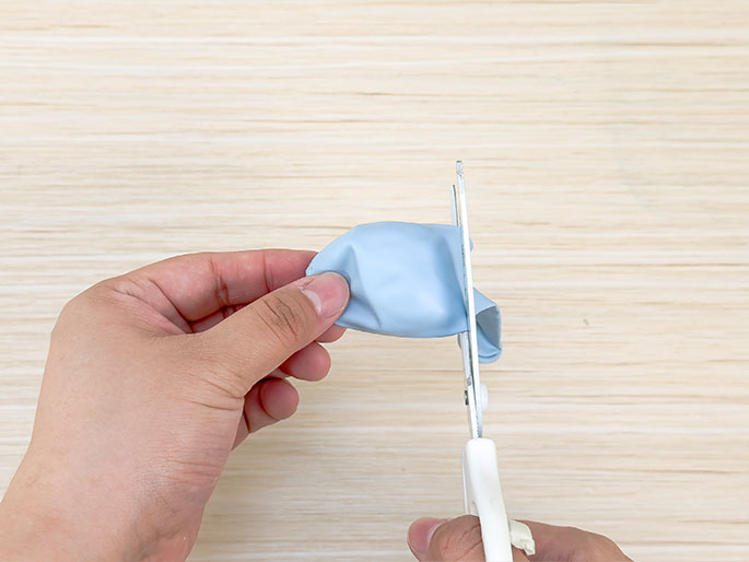 Two hands are using scissors to cut the neck off of a light blue balloon. The balloon is deflated and held taut against a light woodgrain background. This image likely demonstrates a step in a craft or DIY project involving balloons.