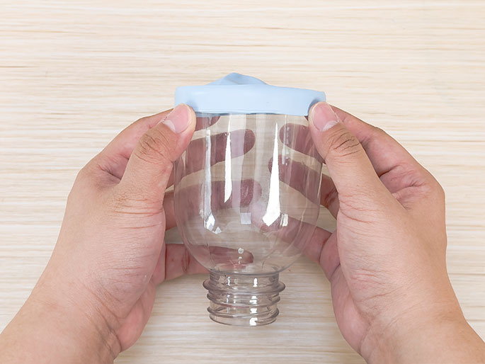 Two hands hold a clear plastic bottle that has a light blue balloon stretched over its opening, creating a seal. The bottle is positioned upright against a light woodgrain background. This image likely shows a stage in a science experiment or DIY project, possibly a homemade air pump or launcher.
