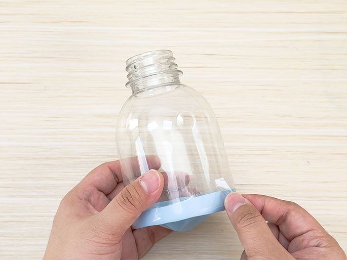 Two hands hold a clear plastic bottle, upside down, with a light blue balloon stretched and secured over the bottom. The balloon is taped around the edges where it meets the plastic. The bottle and balloon are against a light woodgrain background. This appears to be a step in constructing a homemade device, likely for a science project or DIY activity.