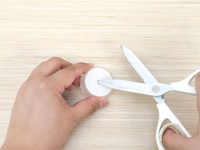Against a light woodgrain background, two hands are shown. One hand holds a white plastic bottle cap, while the other hand uses scissors to make a small cut or hole in the center of the cap. This image likely depicts a step in a DIY project or craft activity involving repurposing a bottle cap.