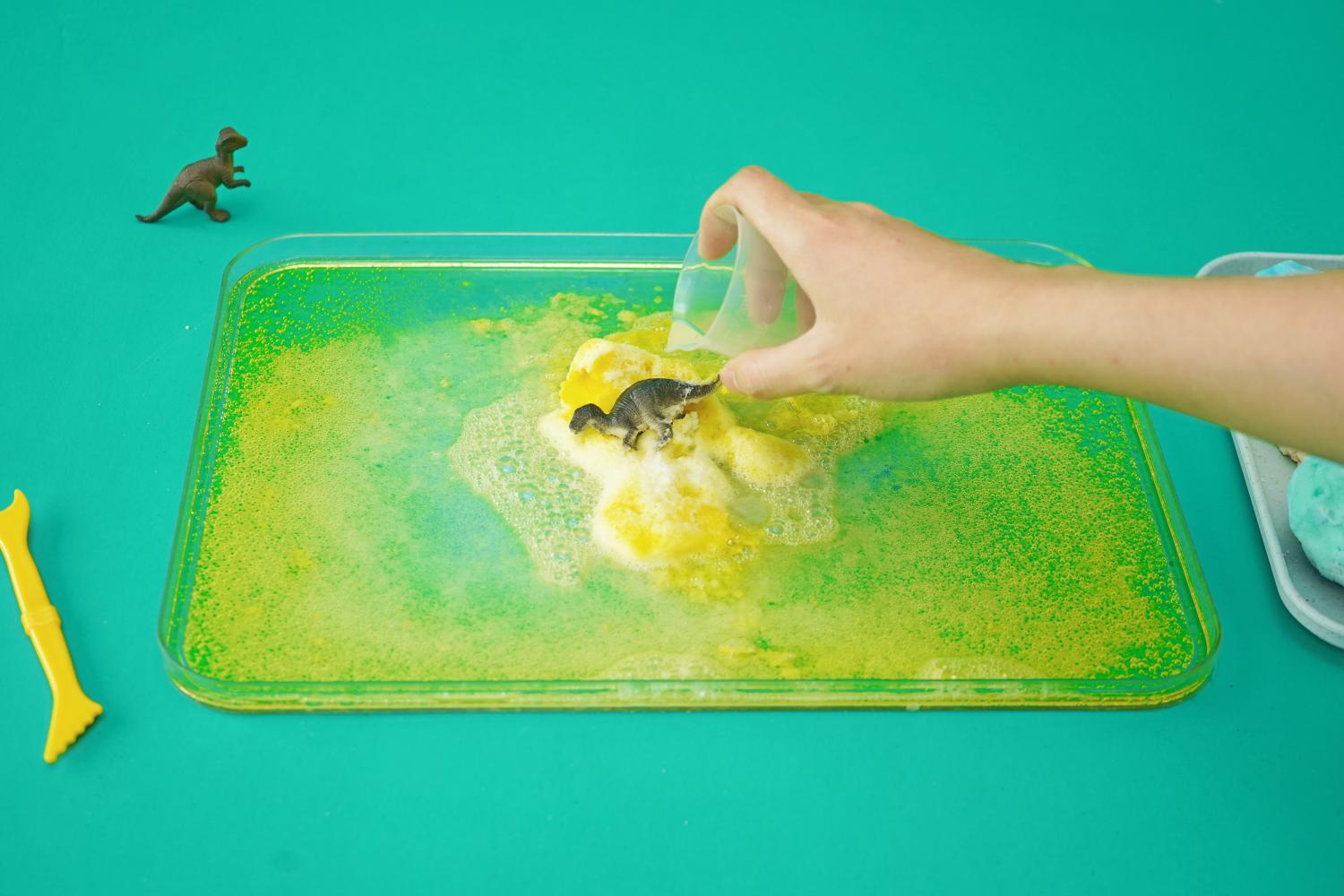 A hand gently places a small, yellow fizzy dinosaur egg into a tray filled with a bubbling, lime-green liquid. A small, dark grey plastic dinosaur is visible in the top left corner, alongside a yellow plastic tool. The egg is partially submerged, and the liquid is visibly fizzing around it, indicating a chemical reaction. The tray is rectangular and clear, allowing a clear view of the reaction. The bright green background provides a strong contrast to the yellow liquid and the egg. The overall image suggests a fun and engaging science experiment in progress.