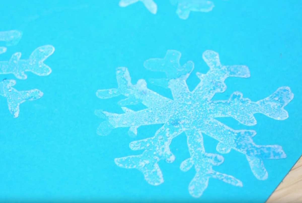 Delicate, glistening snowflakes, seemingly made of ice or a similar translucent material, are scattered across a vivid blue surface. The snowflakes vary slightly in size and shape, capturing the intricate and unique beauty of natural snowflakes. A corner of a light wooden surface is visible in the bottom right.
