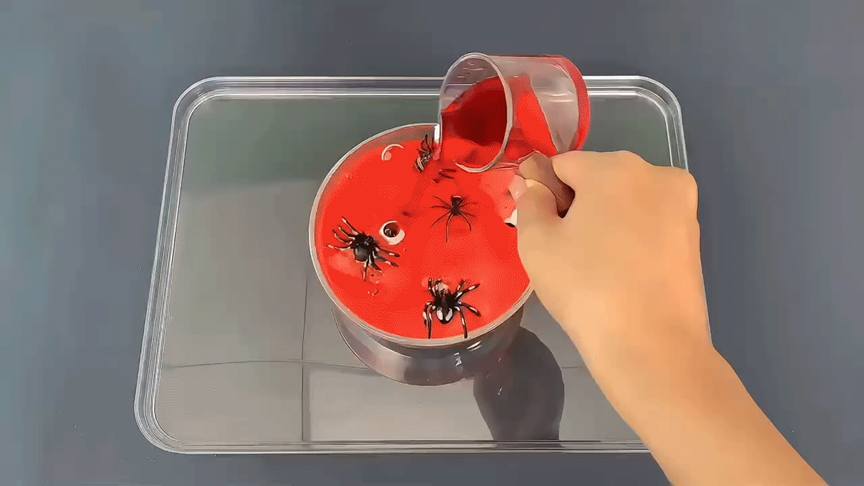 A hand pours a bright red, viscous liquid into a clear, circular container. The container already holds several small, black plastic spiders and a single googly eye, partially submerged in the liquid. The container sits within a larger, clear rectangular tray on a dark grey surface. The red liquid appears thick and almost jelly-like, creating a visually striking contrast against the black spiders and clear containers. The scene suggests a Halloween-themed craft or sensory activity in progress.