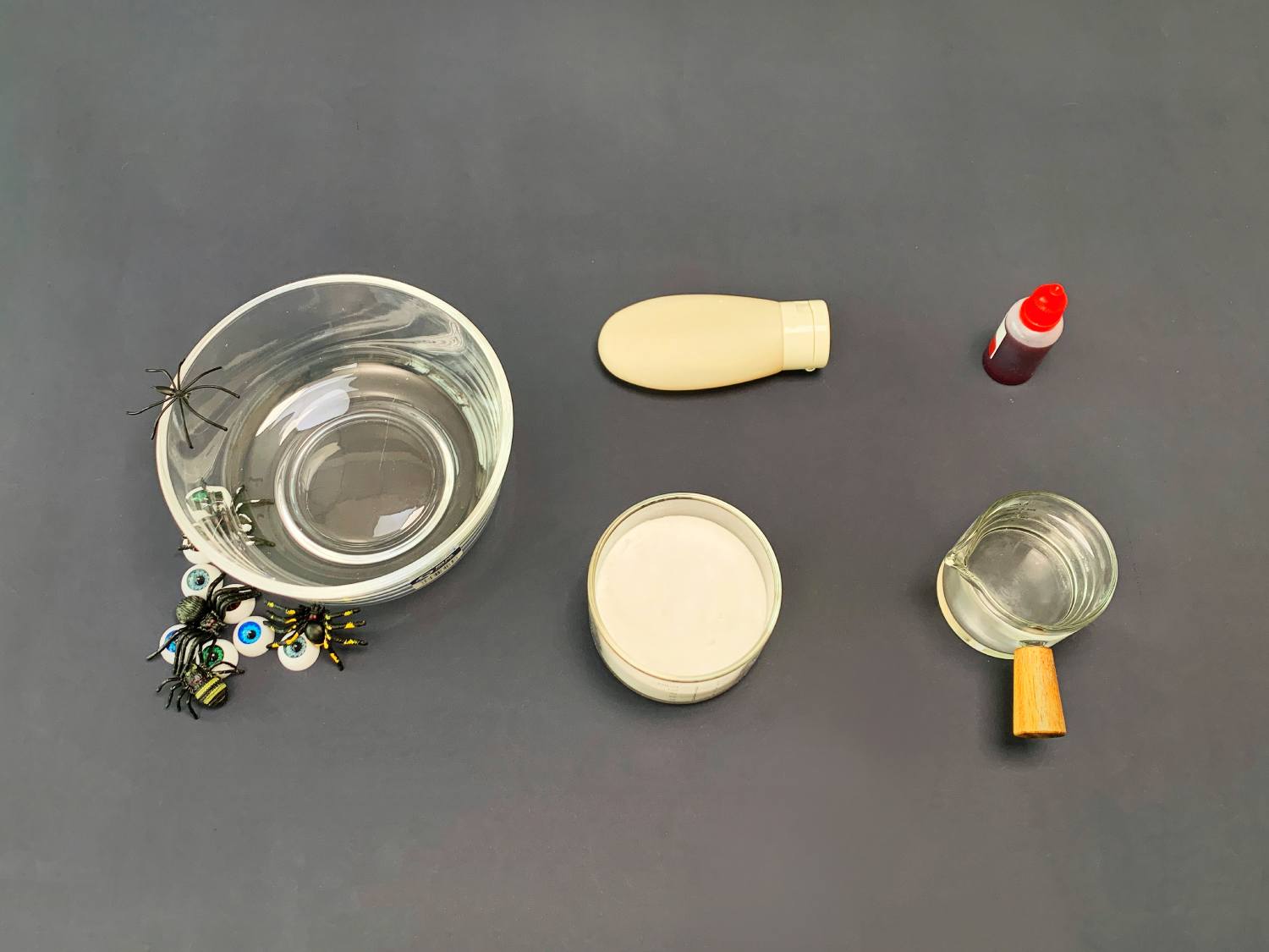 Several items are arranged on a dark grey surface, clearly laid out for a craft project. A glass bowl, decorated with small plastic spiders and googly eyes, sits to the left. Next to it are a small container of what appears to be white powder (possibly borax), a small bottle of red liquid (likely food colouring), and a tube of what looks like white glue or a similar substance. Finally, a small glass measuring jug is positioned on the right. The overall impression is of a preparation for a Halloween-themed slime or similar craft activity.