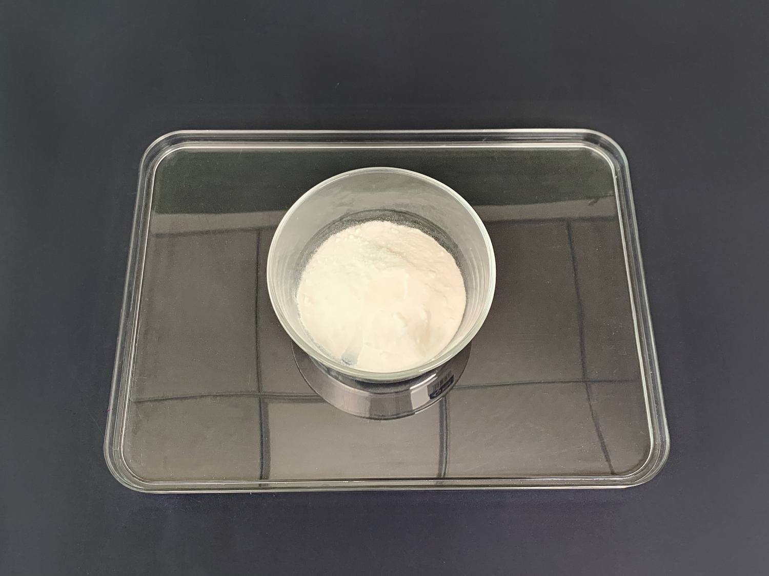 A clear glass bowl containing a quantity of off-white powder sits centrally on a clear plastic tray. The tray rests on a dark grey surface. The powder appears fine and uniformly textured. The image is simple and clearly shows the bowl and its contents, suggesting a preparation stage for a craft or scientific experiment. The lighting is even, and the overall impression is one of cleanliness and precision.