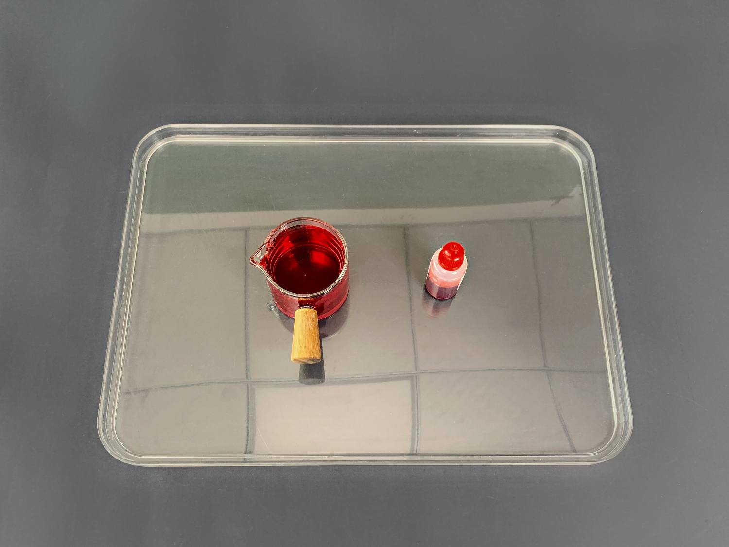 A clear plastic tray sits on a dark grey surface. On the tray are two items: a small, clear glass jug containing a vibrant red liquid and a small, capped bottle of the same red liquid. The jug has a small, light brown wooden handle. The overall impression is of a simple, clean set-up, possibly for a craft or science experiment involving the red liquid, perhaps food colouring. The clear tray and even lighting allow the colours to be clearly visible.