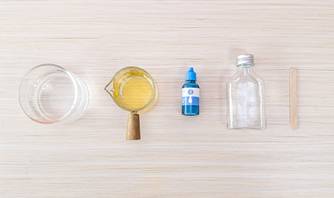 Arranged on a light wooden surface are several items: a glass of water, a small jug of yellow liquid, a bottle of blue colouring, an empty glass bottle, and a wooden stirrer.