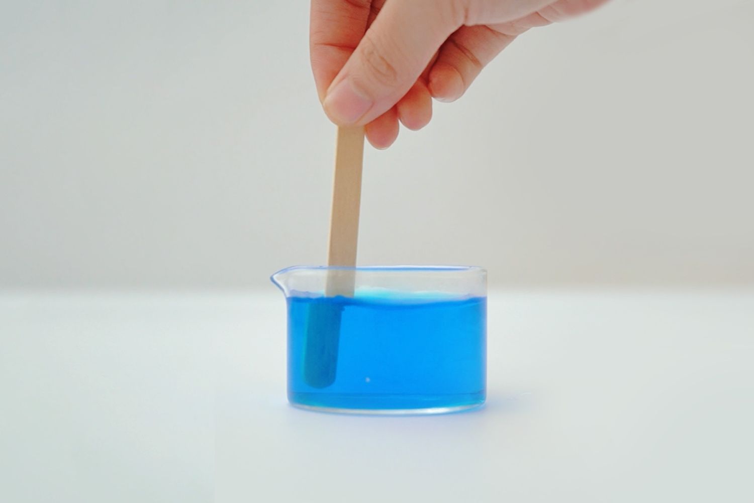 A hand gently holds a small, light-coloured wooden craft stick, partially submerged in a short, cylindrical glass beaker filled with bright blue liquid. The beaker sits on a plain white surface against a similarly light background. The image focuses on the simple act of stirring or mixing the liquid, suggesting a step in a craft or science project, possibly related to creating a colour-mixing sensory bottle. The overall impression is one of calm precision and quiet activity.