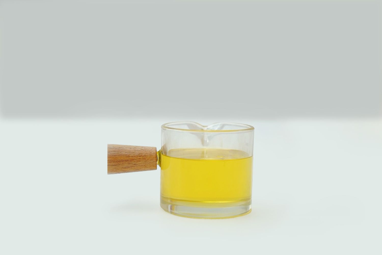 A small, clear glass beaker with a light brown wooden handle sits on a white surface against a pale grey background. The beaker is partially filled with a bright, clear yellow liquid. The liquid appears to be viscous and still, with minimal surface disturbance. The overall impression is one of simplicity and clarity, suggesting a single element in a larger process, perhaps a step in a science experiment or a craft project.