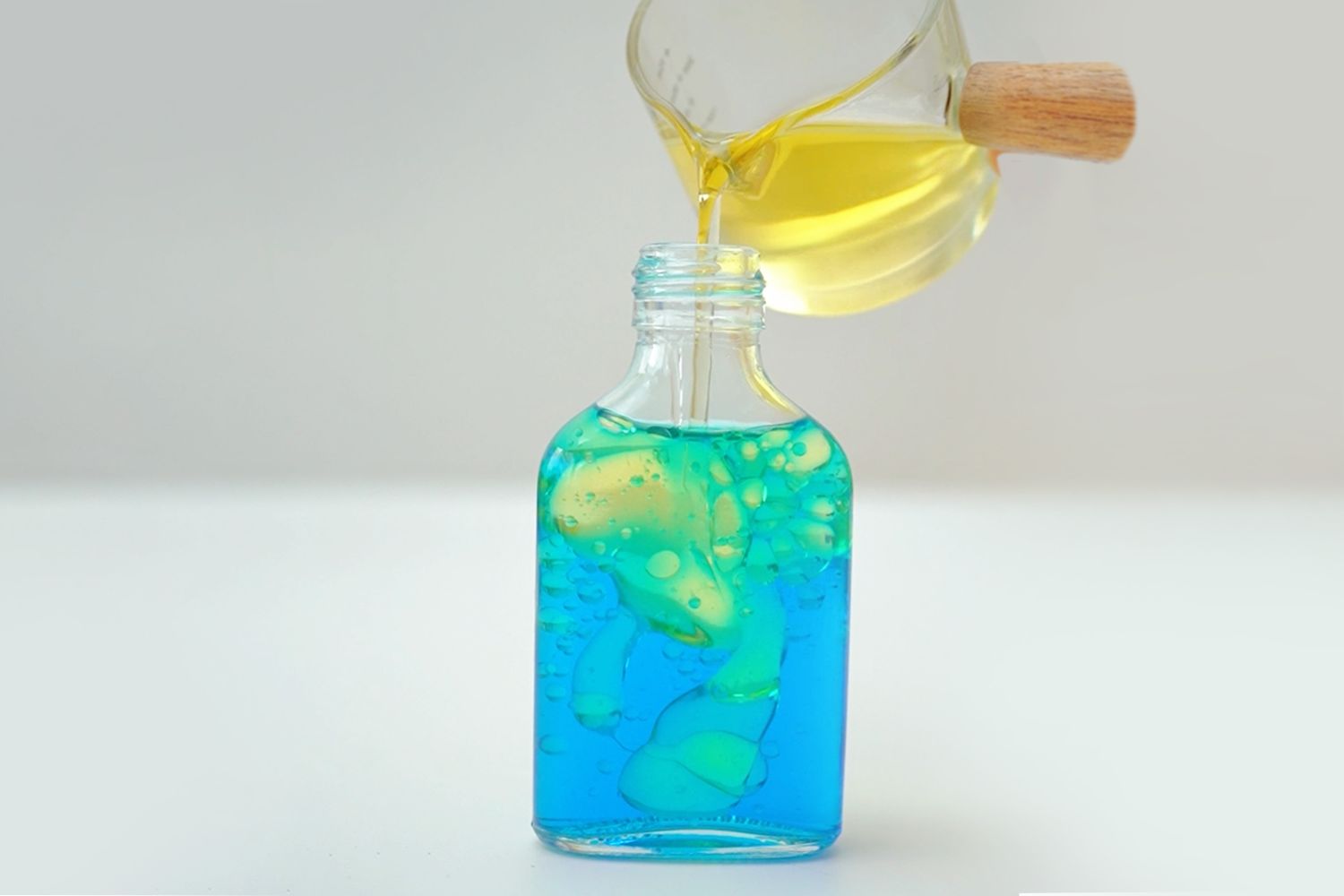 A clear glass jug with a light brown wooden handle is pouring a bright yellow liquid into a small, clear glass bottle. The bottle already contains a layer of bright blue liquid. The yellow liquid is slowly entering the bottle, creating a visible separation between the two liquids and some small bubbles where they meet. The scene is set against a plain white background with a light grey backdrop, creating a clean and simple visual. The overall impression is one of a calm, controlled process, likely part of a craft or science project involving liquid layering.