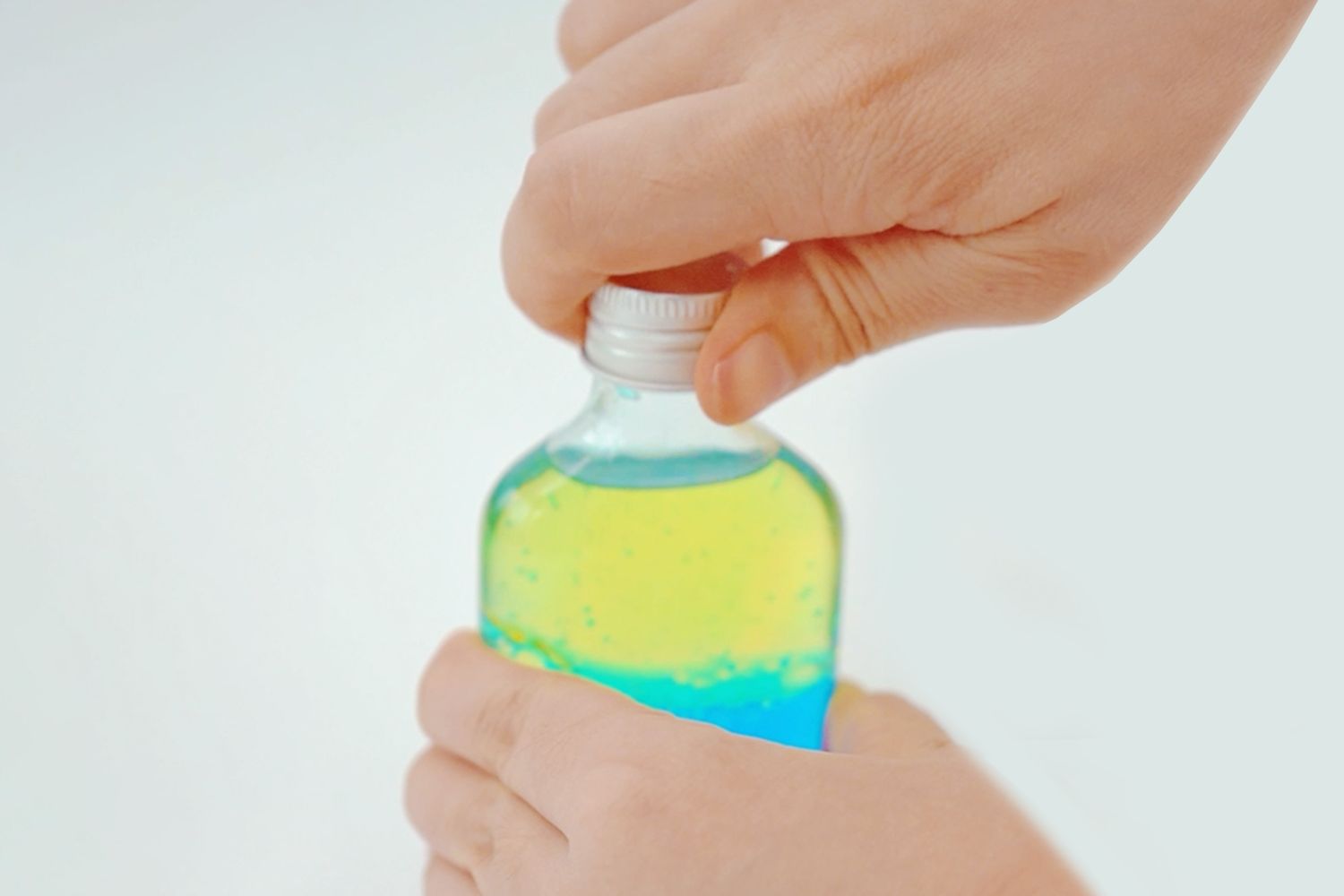 Two hands carefully secure a lid onto a small, clear glass bottle. The bottle contains two distinct layers of liquid: a bright yellow layer on top and a vibrant blue layer underneath. The liquids appear to be slightly viscous, with minimal mixing visible. The bottle is held firmly but gently, suggesting care in sealing the contents. The setting is simple, with a bright, uncluttered background, focusing attention on the bottle and the action of securing the lid. The overall impression is one of a final step in a craft or science project.