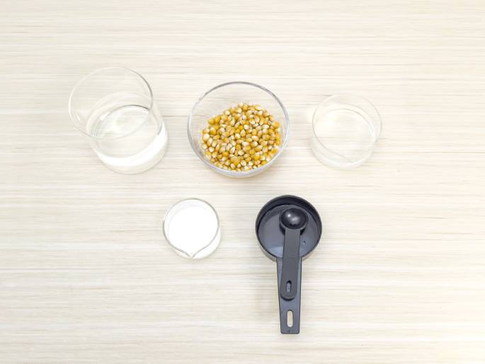 An assortment of ingredients and measuring tools are arranged on a light woodgrain table, suggesting preparation for a recipe. A small glass bowl holds dried corn kernels. Two clear glass beakers, one larger and one smaller, contain clear liquids, likely water. A smaller beaker holds a white liquid, possibly milk. A black measuring cup with a small black measuring spoon inside completes the scene.