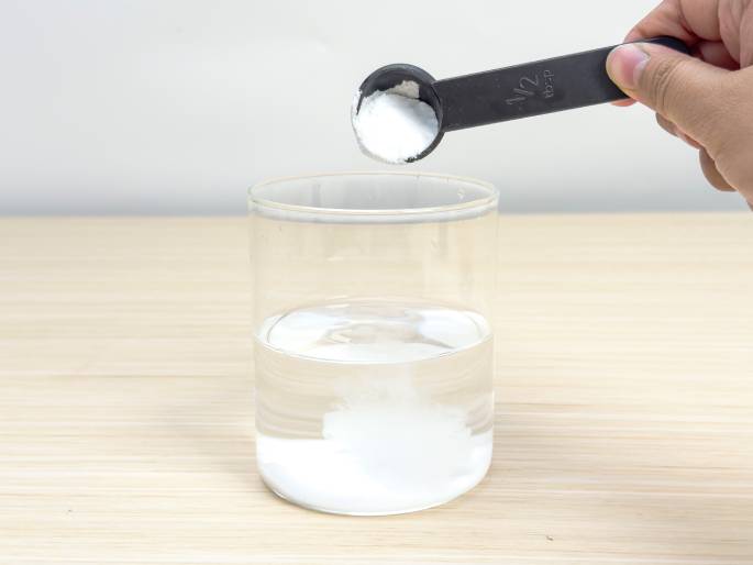 A hand holds a black half-tablespoon measuring spoon filled with white powder over a clear glass beaker containing water. The powder is being poured into the water, creating a small white cloud of dissolving substance at the bottom of the beaker. The beaker sits on a light woodgrain table. The image suggests a science experiment or the mixing of ingredients for a recipe.