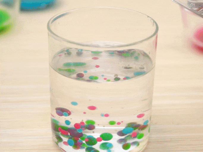 A clear glass sits on a light wooden surface. The glass is partially filled with water and numerous small, colorful orbs. The orbs vary in size and color, with shades of pink, green, blue, and purple visible. More water is poured into the glass from a pitcher, causing the colorful orbs to rise and disperse throughout the water. The video then zooms in for a closer look at the orbs suspended in the liquid, highlighting their vibrant colors and varied sizes. Small air bubbles cling to some of the orbs. The video showcases a visually appealing mixture of water and colorful, buoyant beads or gel balls.