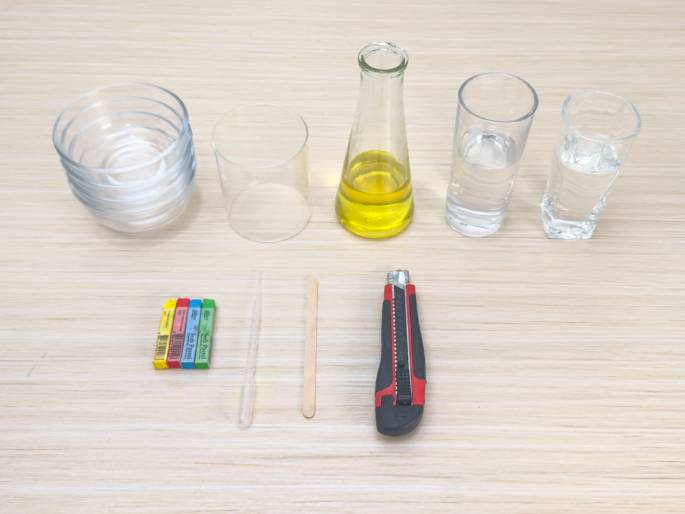 A collection of items arranged on a light woodgrain table suggests preparation for a science experiment or craft project. From left to right, there are: a stack of small clear glass bowls, a larger clear glass cylinder, a small glass bottle containing a yellow liquid (possibly oil), two clear drinking glasses containing water, a small group of colorful wax crayons, a clear plastic pipette or straw, a flat wooden craft stick, and a retractable utility knife with a red handle. The arrangement and variety of items imply a multi-step process.