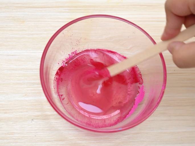 A small, clear glass bowl containing a vibrant pink liquid sits on a light-colored wooden table. A hand holds a small wooden stick, stirring the liquid. Some undissolved pink pigment is visible, suggesting the color is being mixed into the liquid. The scene implies an art project, a science experiment, or the preparation of a cosmetic product.