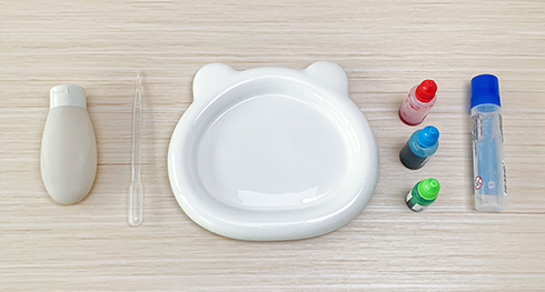 Gather your materials for some fizzy fun! You'll need a small bear-shaped dish, a bottle of clear glue, a pipette, and bottles of red, blue, and green food colouring. Let's get those miniature fireworks ready!
