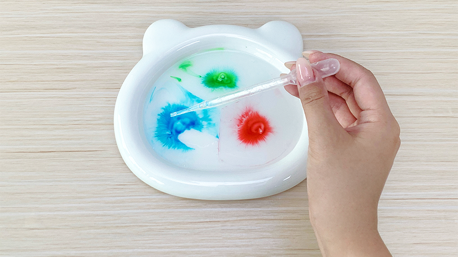 Adding a splash of colour! A hand uses a pipette to carefully add more blue food colouring to the glue in the bear-shaped dish, creating a vibrant and swirling effect. Those colours are really starting to dance!