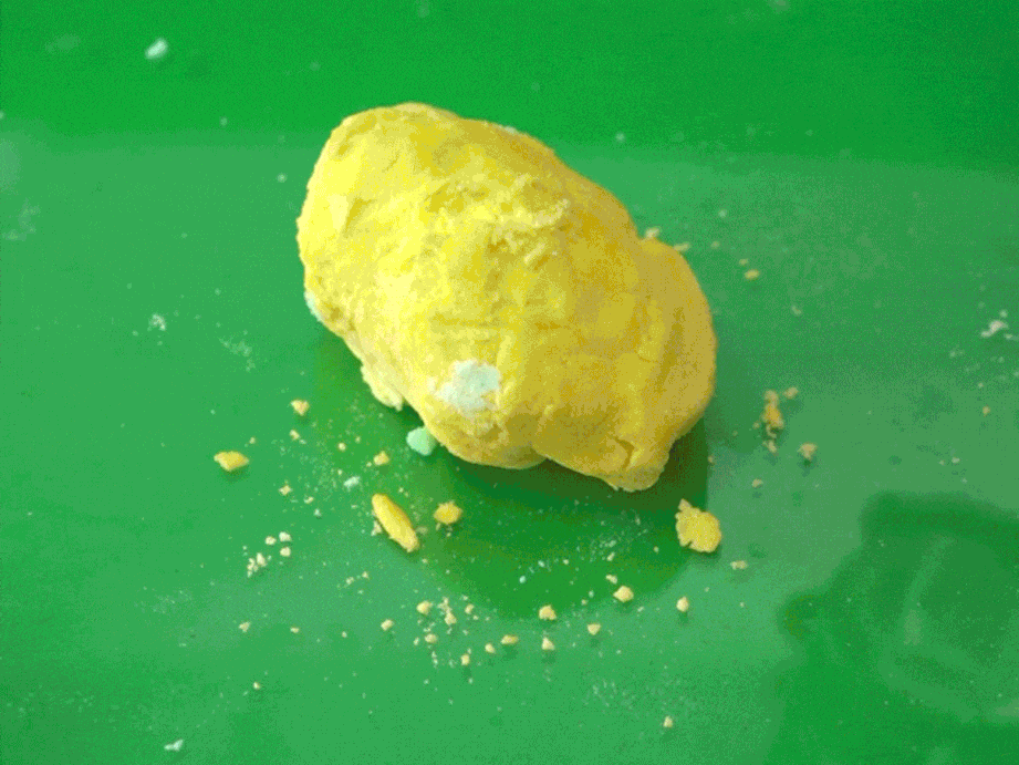 A yellow bath bomb fizzes and dissolves in a tub of water, creating a bubbly, light green solution. As the bath bomb disintegrates, it reveals a small, black plastic scorpion toy hidden inside, adding a playful surprise to the bath time experience.