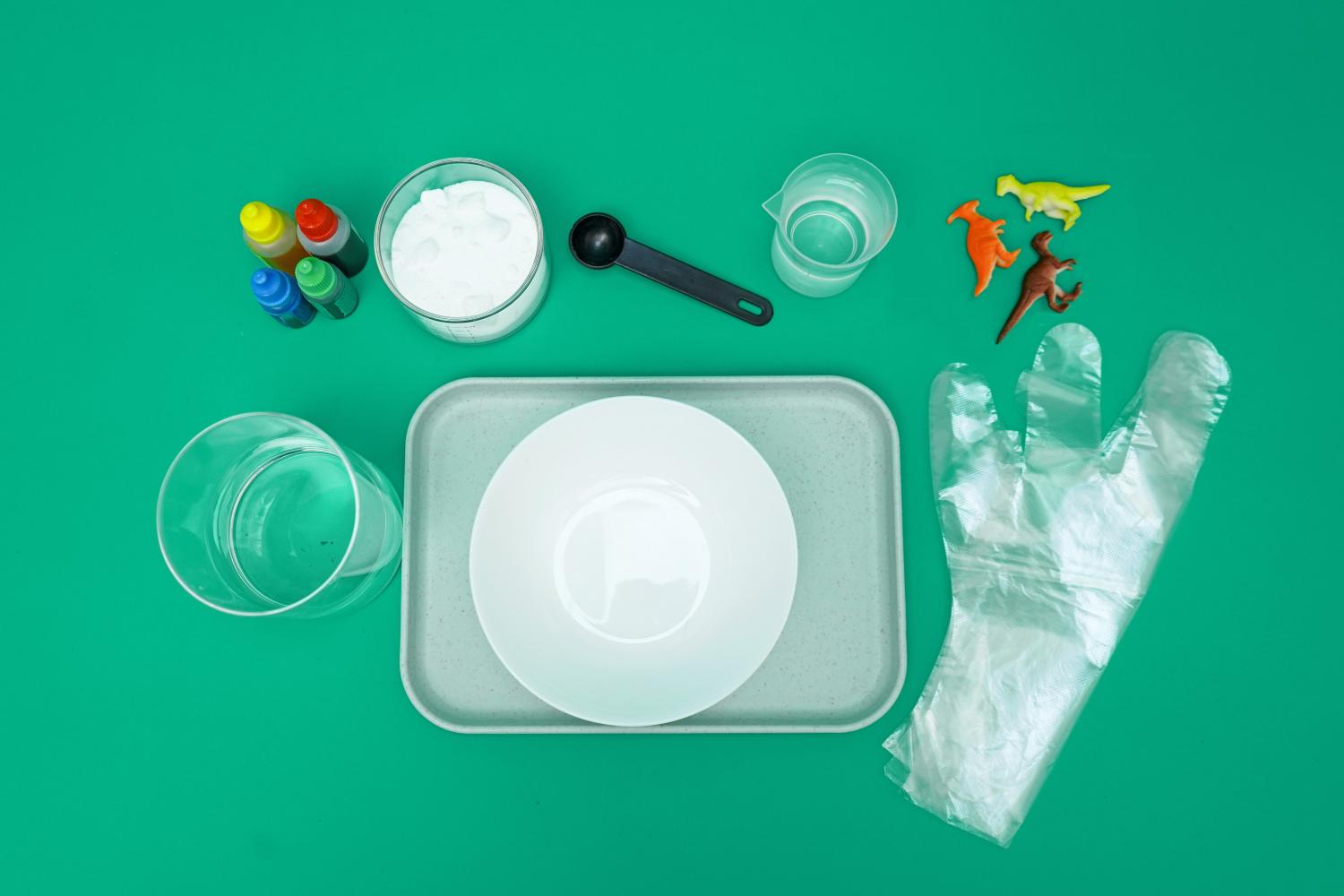Arranged on a vibrant green surface are the ingredients and equipment needed for a fizzy dinosaur egg experiment. These include a small container of baking soda, several small bottles of coloured liquid, a measuring jug containing water, a small measuring cup, a spoon, a clear glass, a white plate on a grey tray, and a plastic glove. Three small plastic dinosaurs are also present, hinting at the experiment's outcome. The items are neatly organised, suggesting a prepared and organised approach to the activity.