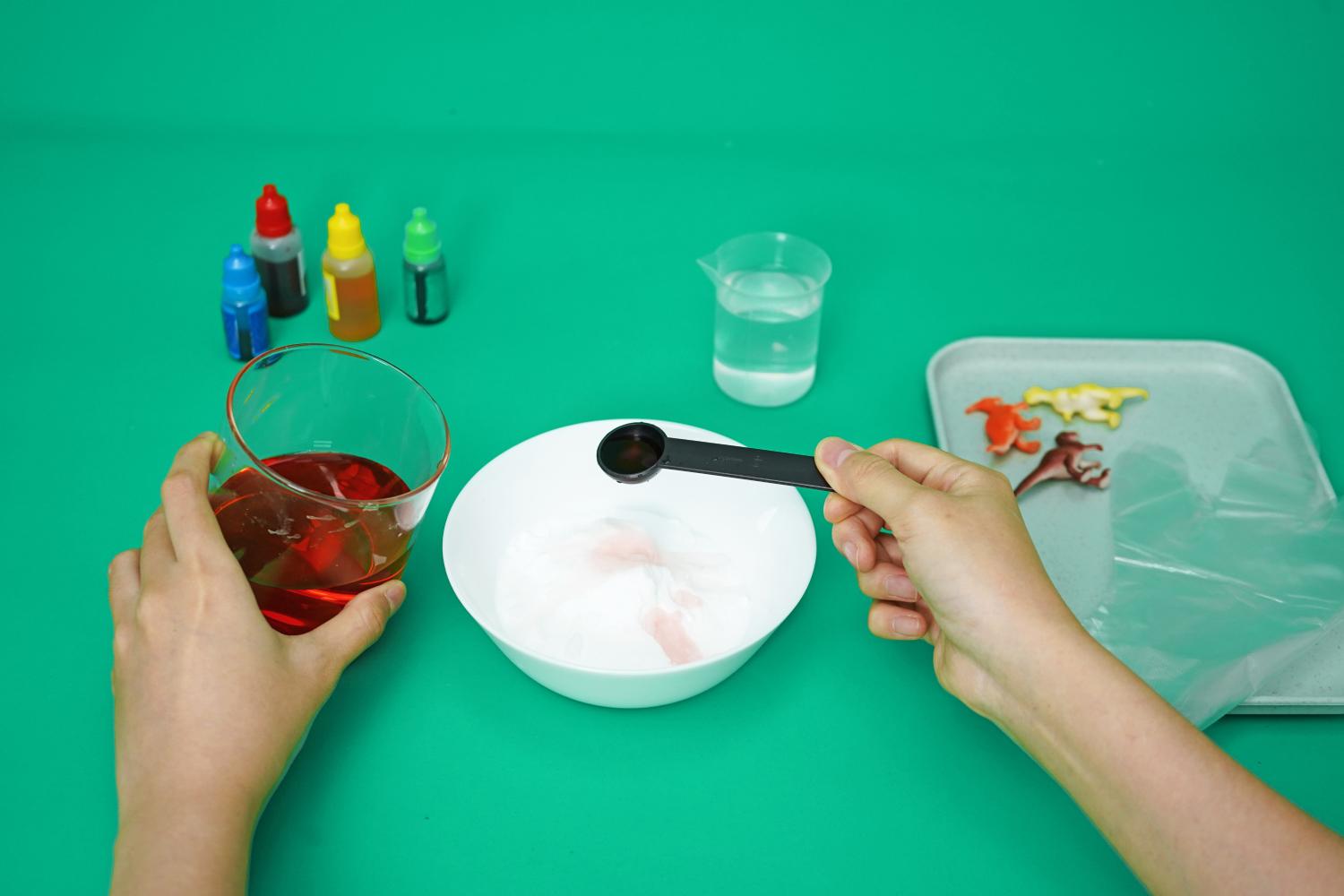 On a bright green surface, a person's hands are shown adding a spoonful of red liquid to a bowl of white baking soda. Several small bottles of coloured liquid are visible in the background, along with a glass of liquid and some small plastic dinosaurs. A grey tray containing a plastic glove is also present. The scene depicts a stage in the preparation of fizzy dinosaur eggs, showcasing the careful addition of coloured liquid to the baking soda mixture. The overall impression is one of a fun and engaging science experiment.