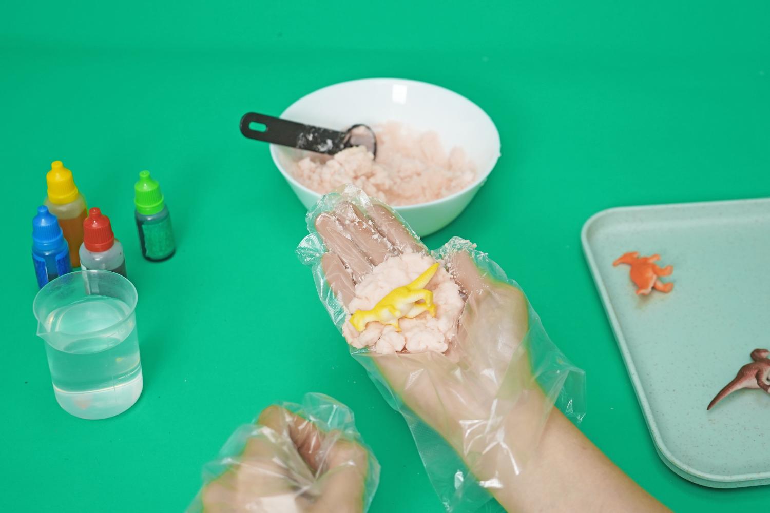 On a bright green surface, a person's hands, wearing disposable gloves, are carefully embedding a small yellow plastic dinosaur into a mound of pink, granular material. A white bowl containing a larger quantity of the same pink mixture is visible in the background, along with small bottles of coloured liquid, a measuring jug of water, and a small tray holding additional plastic dinosaurs. The scene clearly depicts a step in the process of creating fizzy dinosaur eggs, focusing on the careful placement of the dinosaur within the baking soda mixture. The overall impression is one of a fun, hands-on science project.