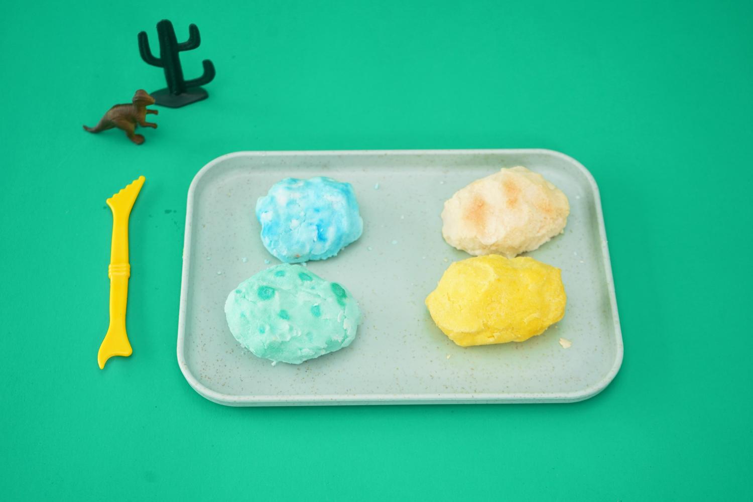 Four colourful, egg-shaped lumps of baking soda mixture rest on a light grey rectangular tray. Two are a pale teal, one is a pale yellow, and one is off-white. They sit on a bright teal background, next to a small brown plastic dinosaur and a small black plastic cactus. A yellow plastic tool, possibly for digging or sculpting, lies to the left. The overall impression is of a completed stage in a children's science experiment, specifically the making of fizzy dinosaur eggs, ready for the next step.