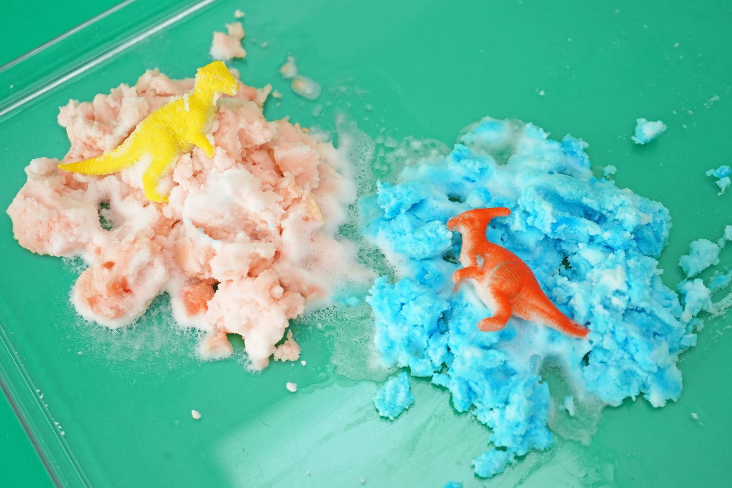 Two fizzy dinosaur eggs, one pink and one blue, have erupted on a clear tray atop a teal surface. A yellow dinosaur emerges from the pink remains, and an orange dinosaur from the blue. The eggs have reacted with vinegar, creating a bubbly, frothy mess. The vibrant colours and the playful dinosaur toys create a visually engaging scene, indicative of a completed, successful science experiment. Bits of the egg mixture are scattered around the main clumps, showing the extent of the reaction.