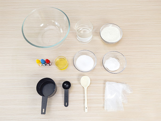 An overhead shot displays various ingredients and tools arranged on a light woodgrain surface, likely for a science experiment or cooking activity. Items include a large clear glass bowl, a glass of water, small bowls containing flour, sugar, and baking soda, three small bottles of food coloring (yellow, blue, and red), a small dish of oil, black measuring cups and spoons, a white spoon, and a small clear plastic bag of an unidentified white substance.