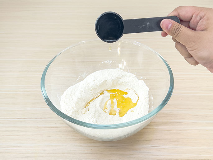 A hand pours oil from a small black measuring spoon into a clear glass bowl containing flour. A small puddle of oil is already visible in the flour. The bowl sits on a light woodgrain surface. This image appears to show a step in a recipe or experiment where oil is being added to dry ingredients.