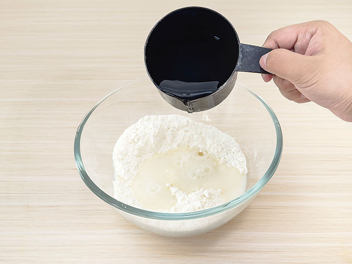 A hand pours a liquid, likely water, from a black measuring cup into a clear glass bowl containing flour. Some of the liquid has already been poured, creating a wet area in the flour. The bowl sits on a light woodgrain surface. This image likely depicts a stage in cooking or an experiment where liquid is being combined with dry ingredients.