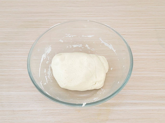 A clear glass bowl sits on a light woodgrain surface. Inside the bowl is a single, cohesive mass of white dough. Residue from the dough preparation is visible on the inside walls of the bowl. This image likely depicts dough that has just been mixed and is ready for the next stage in a recipe.
