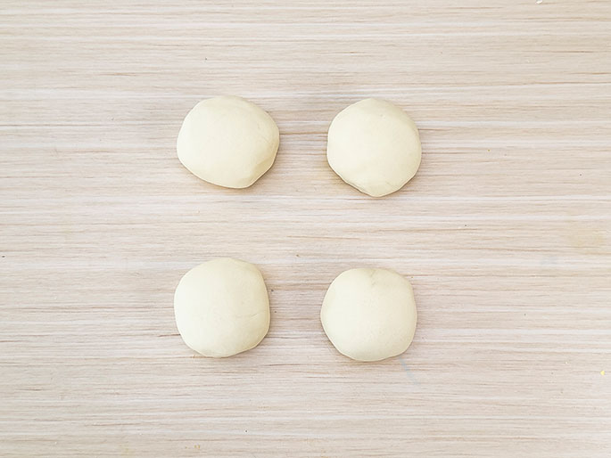 Four equal-sized balls of dough are arranged in a two-by-two grid on a light woodgrain surface. They appear ready to be shaped or cooked.