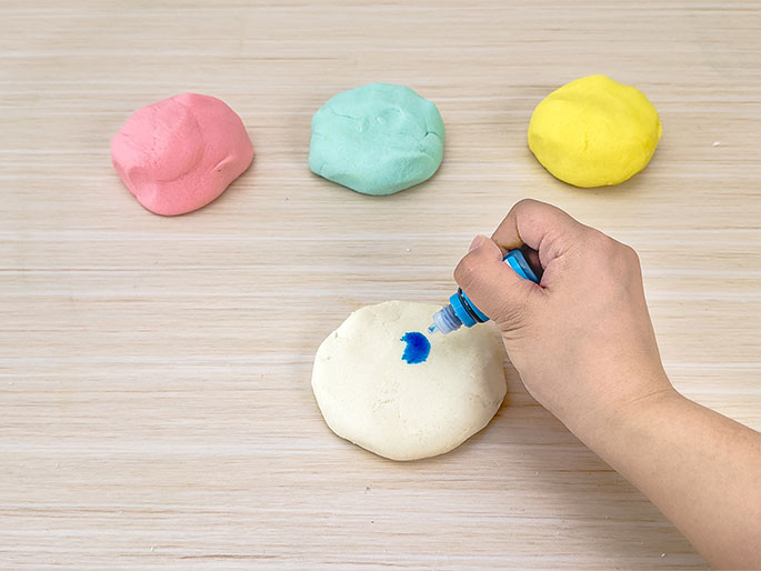 On a light woodgrain surface, a hand uses a small bottle to drop blue food coloring onto a flattened piece of white dough. In the background, three smaller balls of dough are visible: pink, light green, and yellow. This image appears to depict the process of coloring playdough or dough for a craft project.