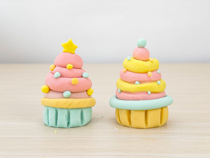 Two small decorative items made of playdough sit on a light woodgrain surface. The item on the left is shaped like a Christmas tree, with layered pink dough forming the tree, decorated with small, colorful dots, a yellow star on top, and a light green base. The item on the right resembles a cupcake or other swirled dessert. It features layered yellow and pink dough swirls, topped with a light green ball, and a light green ring around a textured yellow base. These appear to be children's crafts made from playdough.