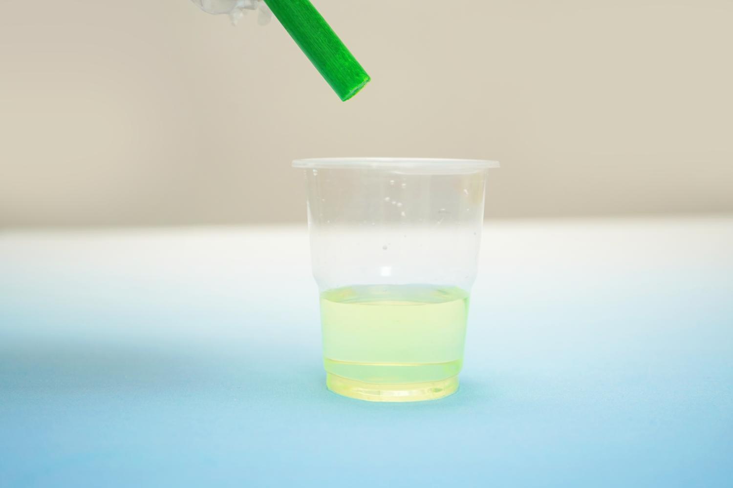 A hand holds a bright green highlighter pen above a clear plastic cup containing a pale yellow liquid. The highlighter is positioned as if its tip is about to be submerged in the liquid, suggesting the process of adding colour. The cup sits on a pale blue surface against a blurred, off-white background. The image is simple and clean, focusing on the action of adding the highlighter to the liquid, possibly for a science experiment or craft project.