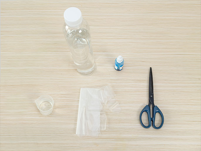 Laid out on a light wood-grain table are several items, seemingly for a craft or science project. These include a clear plastic bottle partially filled with water, a small bottle of blue liquid, a folded piece of clear plastic sheeting, a small clear measuring cup with a small amount of liquid inside, and a pair of blue-handled scissors.