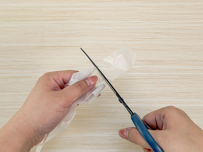 Two hands are shown cutting a piece of clear plastic sheeting with a pair of blue-handled scissors. The sheeting is gathered and held by one hand while the other hand operates the scissors, cutting through the plastic against a light wood-grain background.