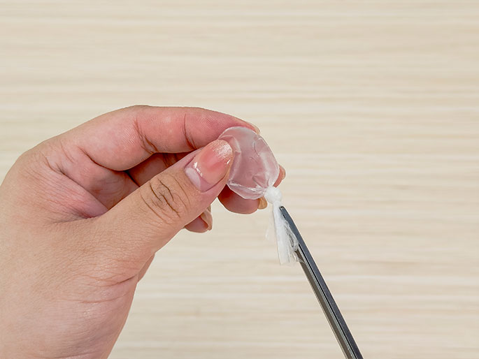 A hand holds a small, clear, water-filled plastic pouch tied at the top. A pair of scissors is positioned to trim the excess plastic just below the knot. The background is a light wood-grain surface.