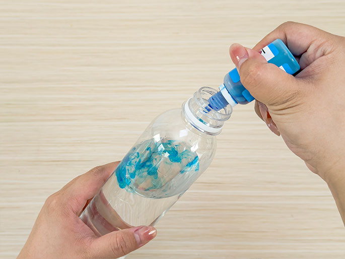 Two hands are adding blue coloring to a clear plastic bottle partially filled with water. One hand holds the bottle while the other squeezes blue dye from a small bottle with a nozzle tip into the water, creating swirls of color. The background is a light wood-grain surface.