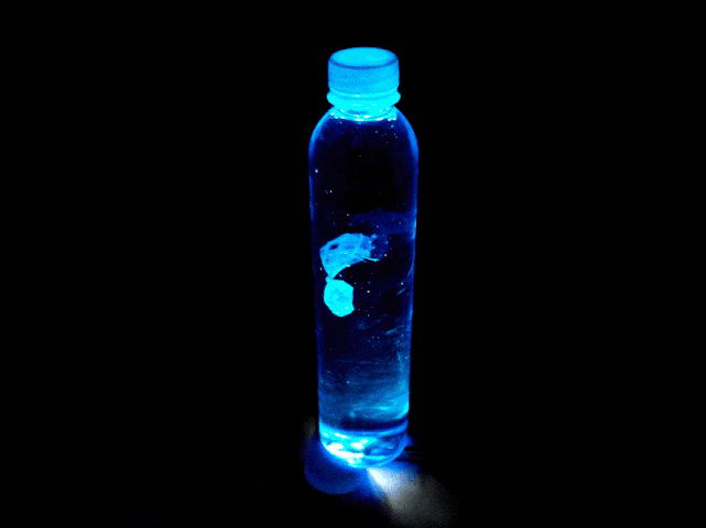 A clear plastic water bottle filled with liquid is illuminated from below by a vibrant blue light against a pitch-black background. A small, jellyfish-like object made of a thin, translucent material drifts and moves gently with the currents of the liquid inside the bottle. The blue light creates a dramatic visual, emphasizing the object and the contents of the bottle.
